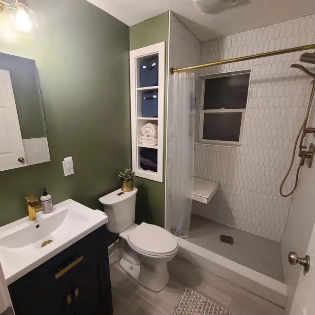 Small bathroom with sage green walls, navy vanity with brass faucet, white picket tile shower with bench and built-in shelves
