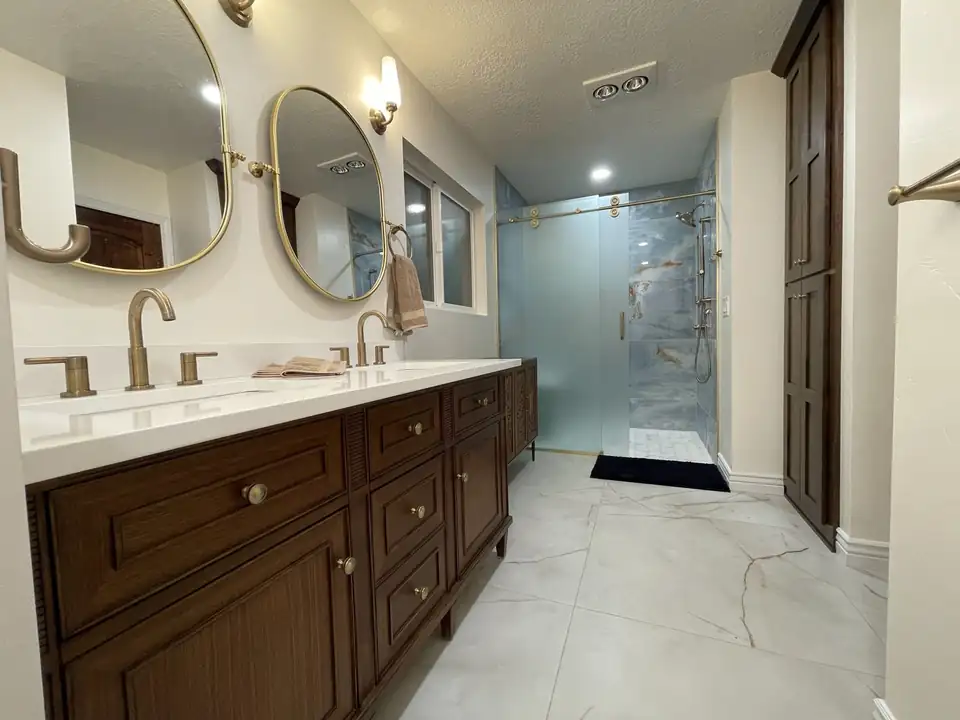 Bathroom remodeling in Utah: master bath with dark wood double vanity, oval brass mirrors, marble-look floor tile and a glass walk-in shower