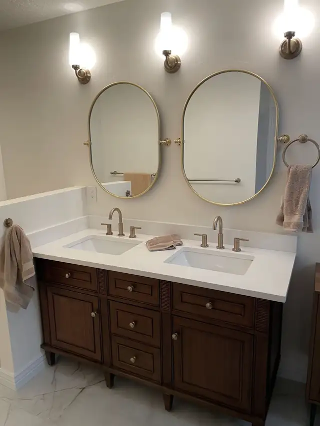 Dark wood double vanity with white top, brushed brass faucets and two oval brass mirrors