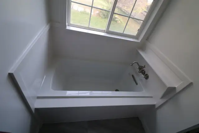 White soaking tub set into a wide ledge beneath a large window, with chrome tub filler